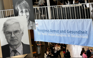 Foto zeigt eine Halle mit zwei Stockwerken, in der Bilder von Personen hängen und ein Banner mit der Aufschrift „Kongress Armut und Gesundheit - Der Public Health-Kongress in Deutschland“. Im Hintergrund sind Personen zu erkennen, die sich miteinander unterhalten.